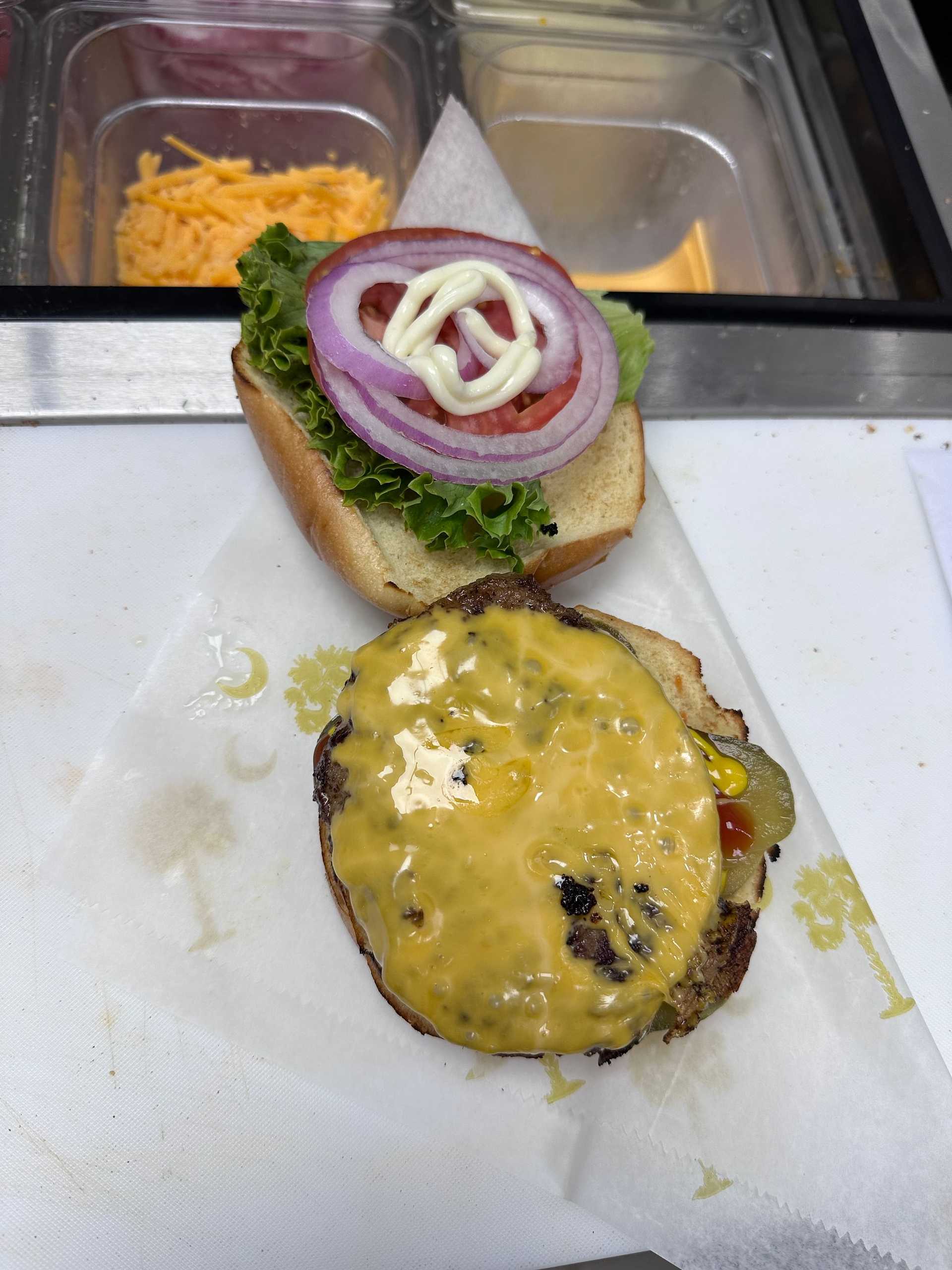 Open cheeseburger being prepared with melted cheese, lettuce, onions, tomato, and mayo on a bun.