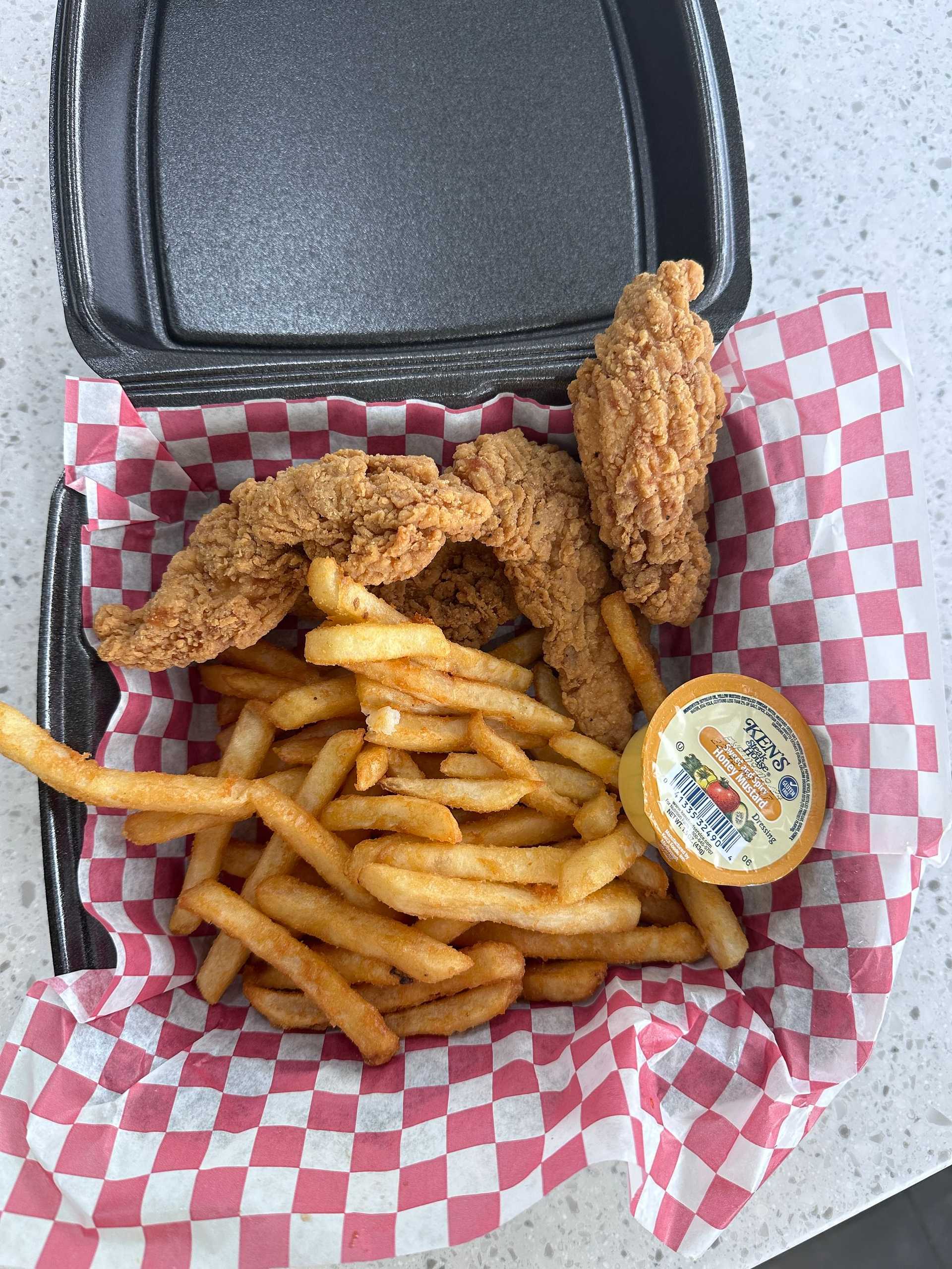 Fried chicken tenders, fries, and honey mustard sauce in a black takeout box on a checkered paper.