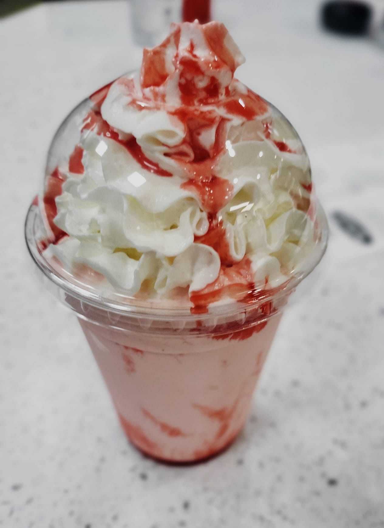 Strawberry milkshake topped with whipped cream and strawberry syrup in a clear plastic cup.